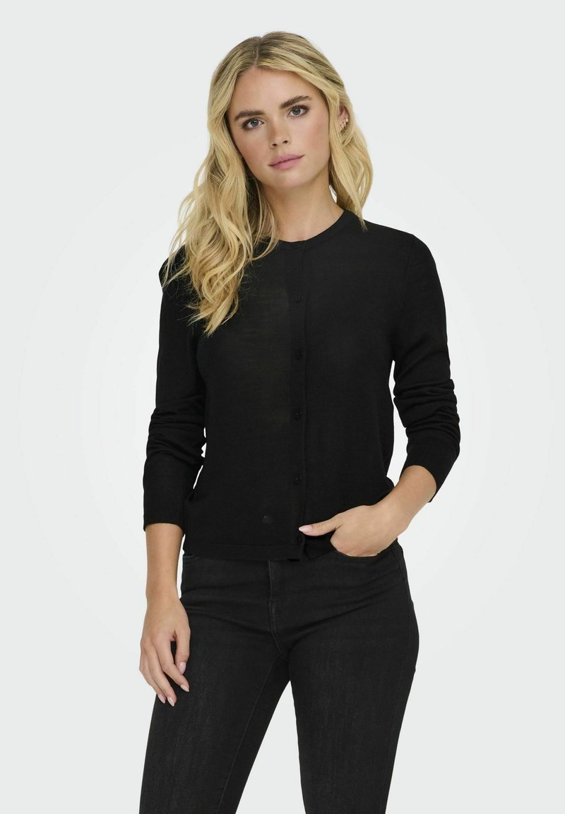 Melna adīta kardigāna ar garām piedurknēm, apaļu kakla izgriezumu un pogu aizdari. Gluda tekstūra, pieguloša silueta, apvienota ar tumšām džinsām.