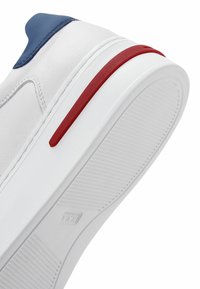 Baskets blanches avec un capuchon de talon bleu et une bande rouge sur la semelle. Elles disposent d'une cuir lisse et d'un fond en caoutchouc texturé avec des stries.