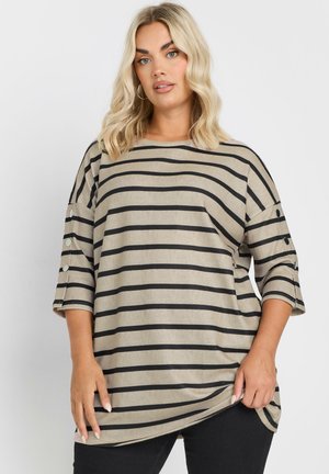 Mujer con cabello rubio hasta los hombros, vestida con una blusa de rayas horizontales beige y negras, con botones en las mangas, sujetando el dobladillo.