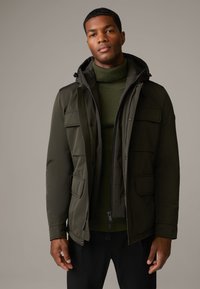 Olivgrüne Jacke mit Reißverschluss auf der Vorderseite und verstellbarem Kapuzenaufsatz, ausgestattet mit mehreren vorderen Taschen und einem strukturierten grünen Pullover darunter.