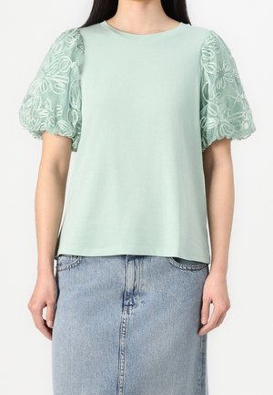 Femme portant un haut vert clair avec des manches bouffantes transparentes brodées et une jupe en jean bleu clair taille haute.