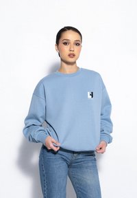 Błękitny sweter z miękkiego materiału, z okrągłym dekoltem, opadłymi ramionami i małym czarno-białym logo na piersi.