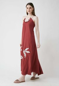 Rotes, ärmelloses Maxikleid mit gerafftem Ausschnitt und weißen Blätterstickereien. Überwiegend entspannte Passform und fransensaum; getragen mit braunen Rutschsandalen.