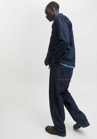 Chaqueta y pantalones de mezclilla azul oscura con un corte relajado. Presenta botones en tono dorado, costuras de doble puntada y una capa de tela más ligera por debajo.