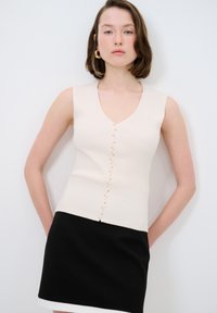Hellbeige gerippter ärmelloser Pullover mit einem V-Ausschnitt und dekorativen Knöpfen auf der Vorderseite, kombiniert mit einem schwarzen Rock mit einem weißen Saum.
