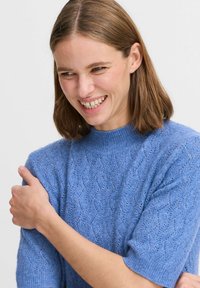 Hellblau gestrickter Pullover mit kurzen Ärmeln, der ein strukturiertes Zickzackmuster und einen gerippten Halsausschnitt aufweist. Das Material wirkt weich und atmungsaktiv.