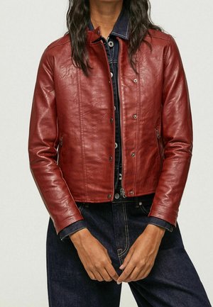 Donna che indossa una giacca di pelle rossa aderente sopra una camicia di denim scuro con bottoni e jeans di denim scuro, con le mani unite in vita.