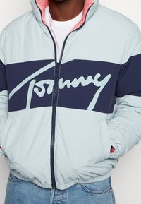 Světle modrá péřová bunda s námořnickou pruhovou výšivkou a velkým bílým logem "Tommy". Růžová podšívka, plný zip a elastické manžety s kapsami.