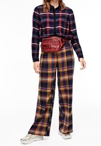 Rutig röd skjorta med knappar, breda rutig byxor i gula, gröna och blå nyanser, parat med en glänsande röd midjeväska och silverskor.