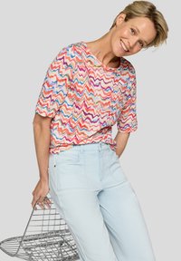 Colorful, short-sleeved top with a zigzag pattern in pink, blue, and green; paired with light blue pants. Holding a wire basket.