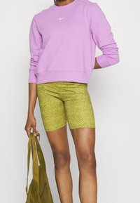 Sweat-shirt violet avec des poignets côtelés, arborant un petit logo blanc, associé à un short cycliste texturé vert et un sac fourre-tout vert.