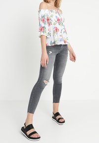 Bílá květinová blůza bez ramínek s růžovými a zelenými vzory, doplněná o šedé džínsy s poškozeným vzhledem a černé sandály s širokými popruhy.