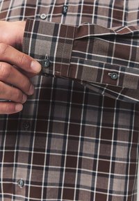 Chemise à carreaux marron et beige à manches longues, avec des boutons foncés et des coutures blanches contrastantes. Détail du poignet avec fermeture par bouton.