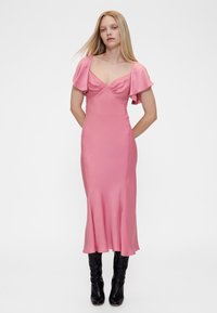 Roza haljina od satena s lepršavim rukavima, uskim gornjim dijelom i proširenim donjim dijelom. Glatka tekstura, dužine do koljena, uparena s crnim čizmama do koljena.