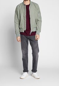 Světle zelená bomber bunda s žebrovanými manžetami, kapsami na zip, vínovým tričkem pod ní, šedými džíny a bílými teniskami.