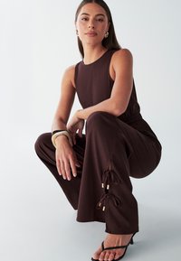 Rudas be rankovių kombinezonas su elastine juosmens dalimi ir raišteliais apačioje. Modelis dėvi juodus sandalus, ant riešo - apyrankė. Neutralios spalvos fonas.