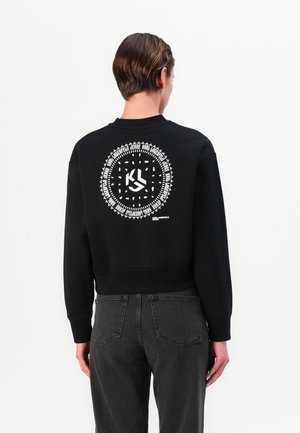 Black cropped sweatshirt with a circular white graphic on the back featuring text and a logo. Ribbed cuffs and hem. Worn with dark gray jeans.