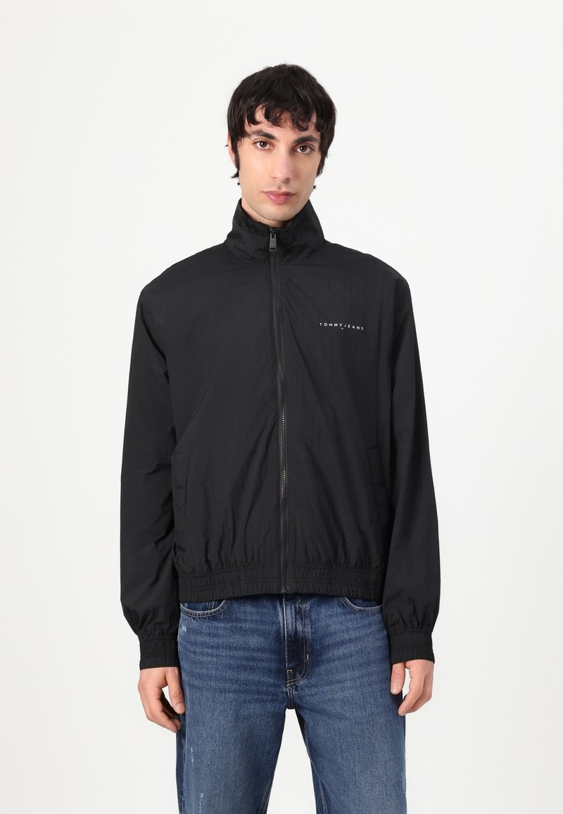 Black lightweight jacket with a high neck, zip closure, elastic waistband, and long sleeves. Features "TOMMY JEANS" logo on the chest.