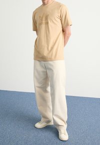 Camiseta de algodón beige con un logo de Adidas texturizado en verde oliva en el pecho, combinada con pantalones de color crema y zapatillas blancas.