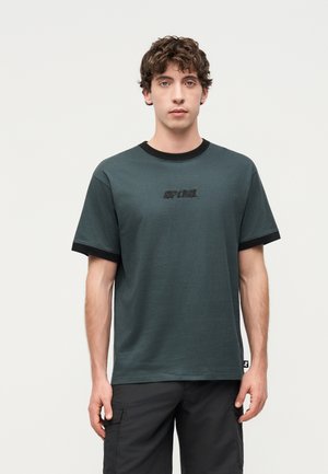 Νεαρός άνδρας με κατσαρά μαλλιά που φοράει σκούρο πράσινο μπλουζάκι Rip Curl με μαύρο γιακά και άκρα μανικιών και μαύρες cargo βερμούδες.