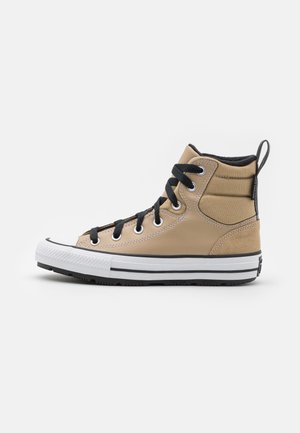 Beige højt sneaker med sorte snørebånd, tyk hvid sål med sorte striber og polstret ankelkrave på hvid baggrund.