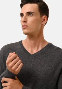 Grau gestrickter Pullover mit V-Ausschnitt-Design. Der Stoff wirkt weich und strukturiert, mit sichtbaren Nähten an den Kanten. Die Hände sind in der Nähe des Ausschnitts positioniert.