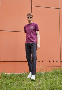 T-shirt in cotone bordeaux con design grafico, abbinata a jeans neri e sneakers bianche, in piedi sull'erba contro uno sfondo di muro arancione.