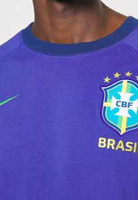 Fialové sportovní tričko s kulatým výstřihem, se zeleným logem Nike a emblémem brazilského národního týmu zdobeným hvězdami a zlatým textem.