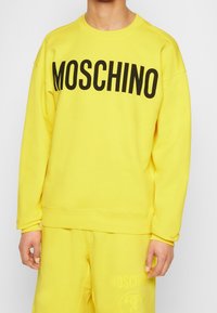 Osoba ubrana w jaskrawożółtą bluzę i spodnie do kompletu, z napisem "MOSCHINO" dużymi czarnymi literami na piersi.