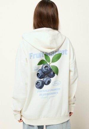Personne portant un sweat à capuche blanc avec un grand motif de myrtille et les textes « Fruit Market » et « Freshly Picked Blueberries » au dos.
