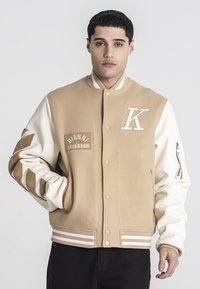 Tamnjakvarsity jakna s kremastim rukavima, s rebrastim prugastim donjim dijelom, izvezenim slovom "K" i oznakom "Gianni Kavanagh" na prsima.