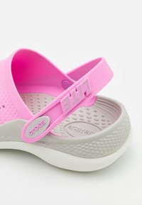 Růžové Crocs s texturovaným vrškem, elastickým páskem na patě a šedou podrážkou. Obsahují značení "crocs" a tvarované tečky pro pohodlí.