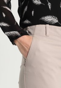 Beige trousers with side pockets and a classic fit, featuring a detailed stitching and a tailored waistband. The top has a black sheer fabric with white feather patterns.