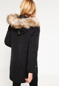 Černá parka s kapucí lemovanou umělým kožíškem, se zipem, zapínáním na patentky a bočními kapsami. Hladký materiál s mírně lesklým vzhledem.