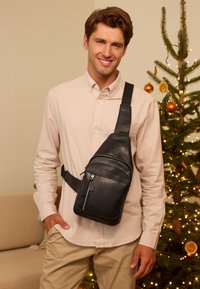 Schwarze Leder-Umhängetasche mit mehreren Reißverschlussfächern, flachem Design und verstellbarem Riemen, der quer über die Brust getragen wird. Im Hintergrund ist ein geschmückter Baum zu sehen.