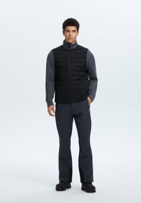 Chaleco acolchado negro con cuello alto sobre una camisa de manga larga gris, acompañado de pantalones gris oscuro y botas negras. Texturas suaves, diseño minimalista.