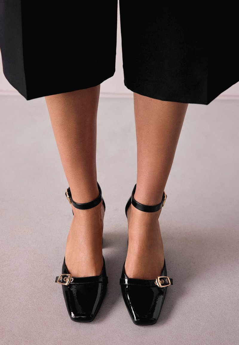 Black patent leather heels with a square toe, ankle strap, and gold buckle detail. Textured surface, elegant design, and a glossy finish.