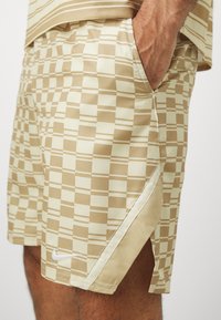 Shorts de sport à carreaux beiges avec fente latérale et logo blanc Nike, portés avec une main dans la poche et un haut rayé beige.