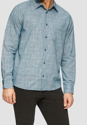 Homme portant une chemise bleue à motifs à manches longues boutonnée et un pantalon foncé, debout avec les mains détendues sur les côtés.