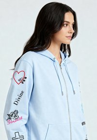Felpa con cappuccio azzurro chiaro con zip, dotata di cappuccio, tasche frontali e ricami sulle maniche, tra cui un cuore e vari testi e icone.