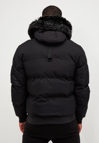 Černá péřová bunda s umělým kožíškem na kapuci, prošívaná textura, elastické manžety a hladký povrch. Obsahuje závěsnou smyčku na zadní straně.