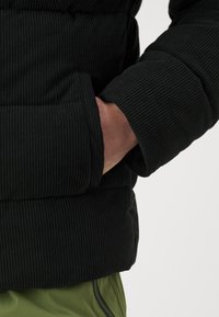 Schwarze Cord-Puffjacke mit gerippter Textur, elastischen Bündchen und einer Seitentasche, kombiniert mit olivgrünen Hosen.