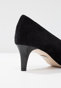 Pompe à talon haut en daim noir avec un talon effilé, une texture lisse et un capuchon de talon noir brillant avec de subtils détails métalliques.