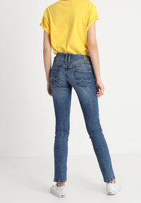 Jeans in denim blu con una vestibilità slim e un'altezza media, dotati di due tasche posteriori con cuciture decorate, abbinati a una t-shirt gialla.