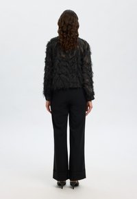 Black feather-textured cropped jacket with sheer fabric, paired with fitted black trousers and pointed black heels, viewed from the back.
