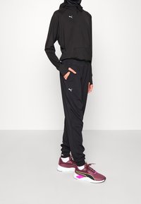 Schwarze sportliche Kapuzenjacke mit einer Fronttasche, kombiniert mit schwarzen Jogginghosen mit seitlichen Taschen. Burgunderfarbene Sneakers mit pinken und gelben Akzenten.