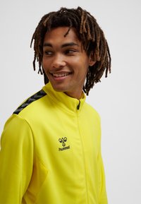 Gul atletisk jakke med lynlåskrave, tekstureret stof og sort logo. Har sorte accenter på skuldrene og sidesømme.