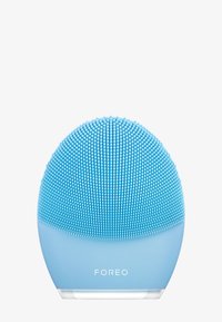 Nettoyant facial en silicone, forme ronde, couleur bleu clair, surface texturée avec de petites poils en silicone, base transparente avec la marque "FOREO".