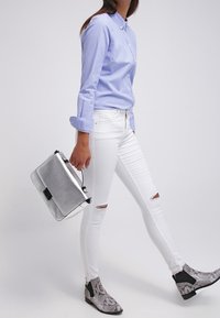 Camicia azzurra con bottoni, jeans skinny bianchi strappati, borsa a tracolla metallica argento e stivaletti alla caviglia grigi con motivo a serpente. Texture morbide e colori a contrasto.