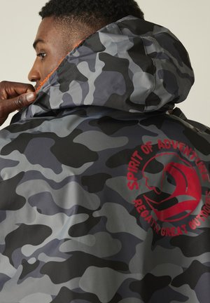 Man met het capuchon van een grijs-zwart camouflagejack met rood logo "Spirit of Adventure Regatta Great Outdoors" op de achterkant aan het aanpassen.